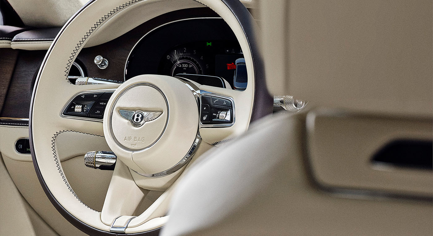 Close-up of a luxury Bentley Spur car interior featuring a beige steering wheel with a logo, intricate stitching, and sleek dashboard controls, exuding elegance.